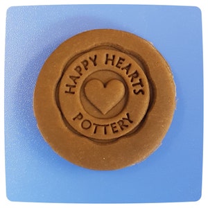 May include: A circular brown clay stamp featuring a central heart icon surrounded by the embossed text HAPPY HEARTS POTTERY. The stamp has a raised circular border and is positioned on a flat, light blue square surface with rounded corners.