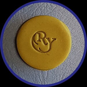 May include: A circular, mustard-yellow wax seal featuring the embossed letters Ry in a serif font. The seal has a smooth, flat surface with a slightly raised edge, centered within a dark blue circular border against a textured gray background.