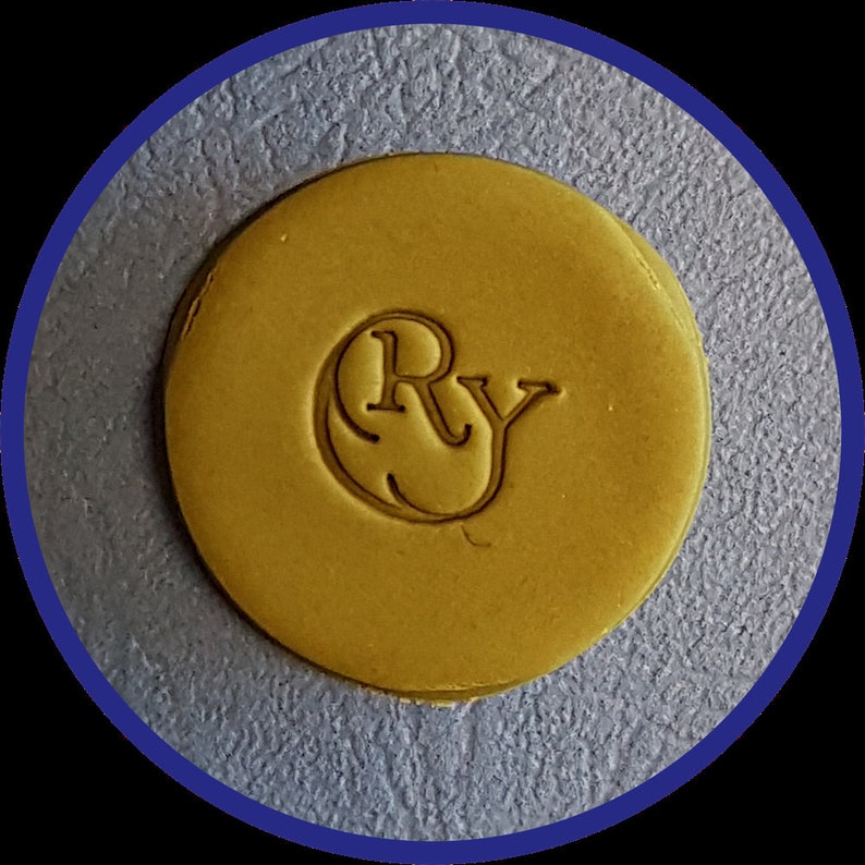May include: A circular, mustard-yellow wax seal featuring the embossed letters Ry in a serif font. The seal has a smooth, flat surface with a slightly raised edge, centered within a dark blue circular border against a textured gray background.