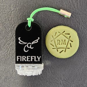 Pode incluir: Um chaveiro preto com um design de libélula branca e a palavra "FIREFLY" impressa nele. O chaveiro tem um cordão verde e um fecho de metal prateado. Também é mostrado um sabonete redondo verde com as letras "RM" no centro.