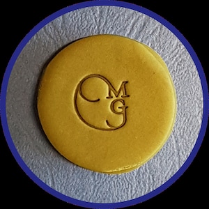 May include: A circular, mustard-yellow wax seal featuring the embossed letters MG inside a stylized C-shaped border. The surface of the wax is smooth and flat, with the letters and circular outline clearly pressed into the material.