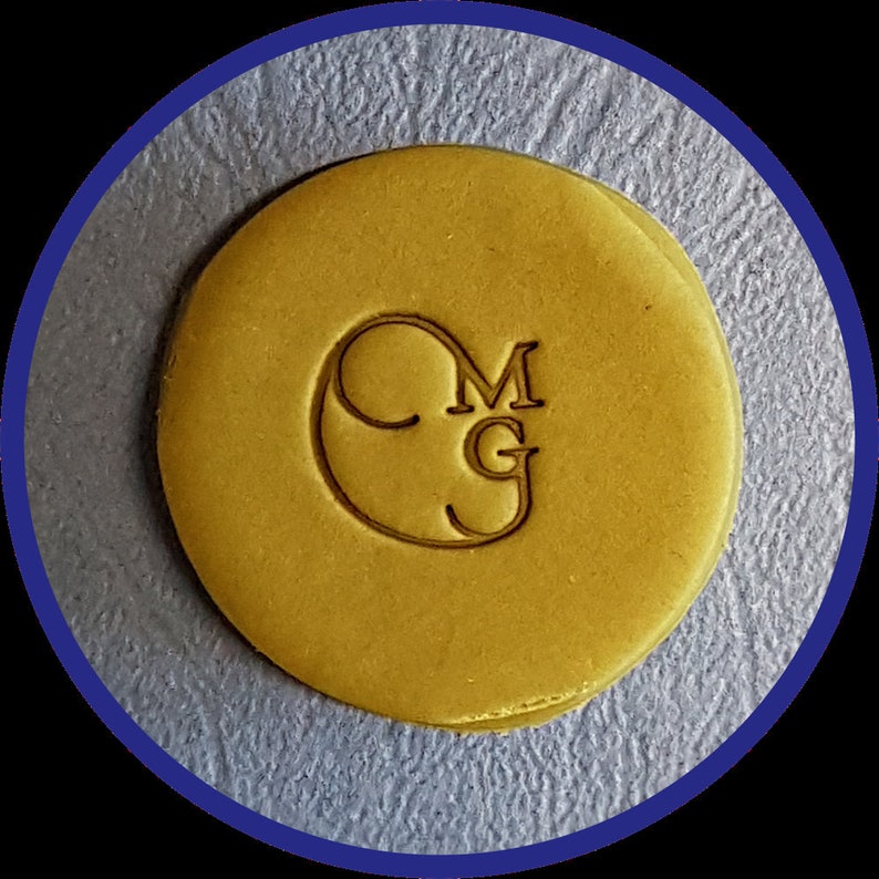 May include: A circular, mustard-yellow wax seal featuring the embossed letters MG inside a stylized C-shaped border. The surface of the wax is smooth and flat, with the letters and circular outline clearly pressed into the material.