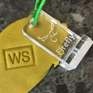 May include: A clear acrylic stamp with the text "Firefly" and a stylized butterfly design. The stamp is being used to imprint the letters "WS" into a yellow dough.