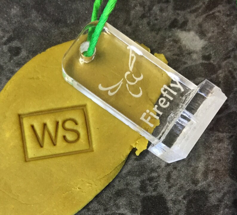 May include: A clear acrylic stamp with the text "Firefly" and a stylized butterfly design. The stamp is being used to imprint the letters "WS" into a yellow dough.
