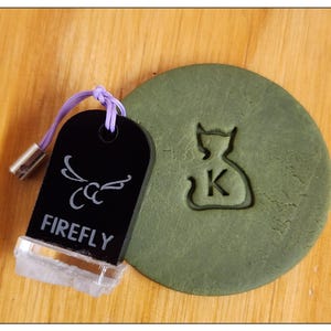 May include: A black and white stamp with the word "FIREFLY" and a stylised design of a dragonfly. The stamp is being used to create an impression of a cat-shaped money bag with the letter "K" inside on a green surface.