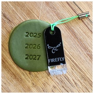 May include: A green round charm with the years 2025, 2026, and 2027 embossed on it. The charm is attached to a black tag with the word "FIREFLY" and a stylised insect design. The tag is attached to a green string with a silver metal clasp.