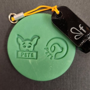 May include: A round green silicone stamp featuring two embossed designs: a cartoon animal head above a rectangular box with the text PETE, and a circular logo with the text PETE. An orange cord attaches a black acrylic tag with a white logo.