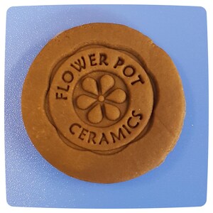 Pode incluir: Um disco de argila marrom com um carimbo circular que diz "Flower Pot Ceramics" com um desenho de flor estilizado no centro.