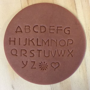 May include: A brown clay stamp with the alphabet A-Z, a sun symbol, and a heart symbol.