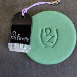 May include: A black acrylic stamp with the word firefly and a butterfly icon printed in white, attached to a light purple cord. Next to it is a round, light green clay disc featuring an embossed logo with the letters DZ inside a horseshoe shape.