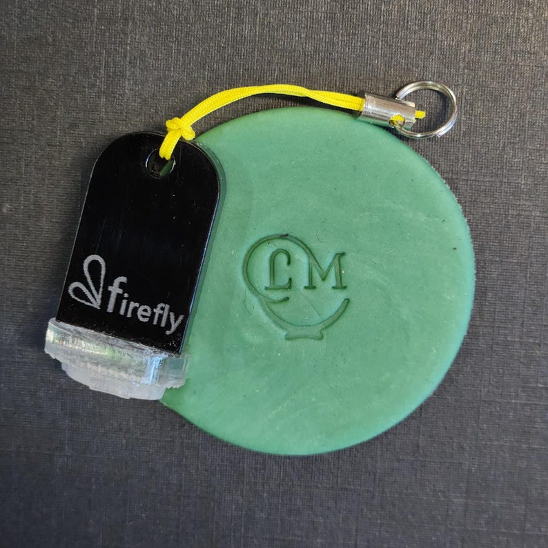 May include: A round green wax seal with an embossed CM logo, attached by a bright yellow cord to a black rectangular tag featuring the white text firefly and a small icon. The tag has a clear base and a silver metal ring for attachment.