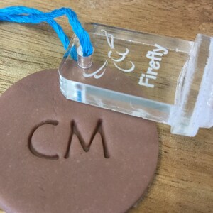 May include: A clear plastic stamp with the text "Firefly" and a decorative design. The stamp is being used to imprint the letters "CM" into a brown clay disc.