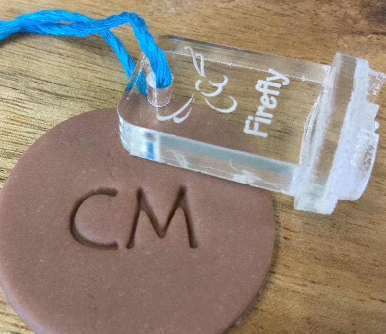 May include: A clear plastic stamp with the text "Firefly" and a decorative design. The stamp is being used to imprint the letters "CM" into a brown clay disc.