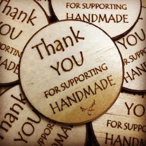 May include: Close-up of several round wooden tags with the text "Thank You" and "For Supporting Handmade" engraved on them. The tags are stacked on top of each other.