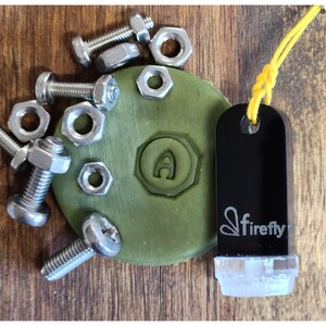 Pode incluir: Uma variedade de parafusos e porcas de metal, e um objeto verde circular com a letra "A" em relevo. Uma etiqueta preta com a palavra "firefly" e um cordão amarelo também estão presentes. Sobre uma superfície de madeira.