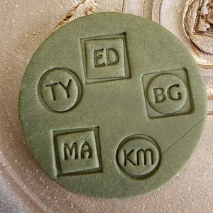 Pode incluir: Um carimbo circular verde com cinco combinações de letras diferentes: TY, ED, MA, KM e BG. As letras estão em relevo no carimbo.