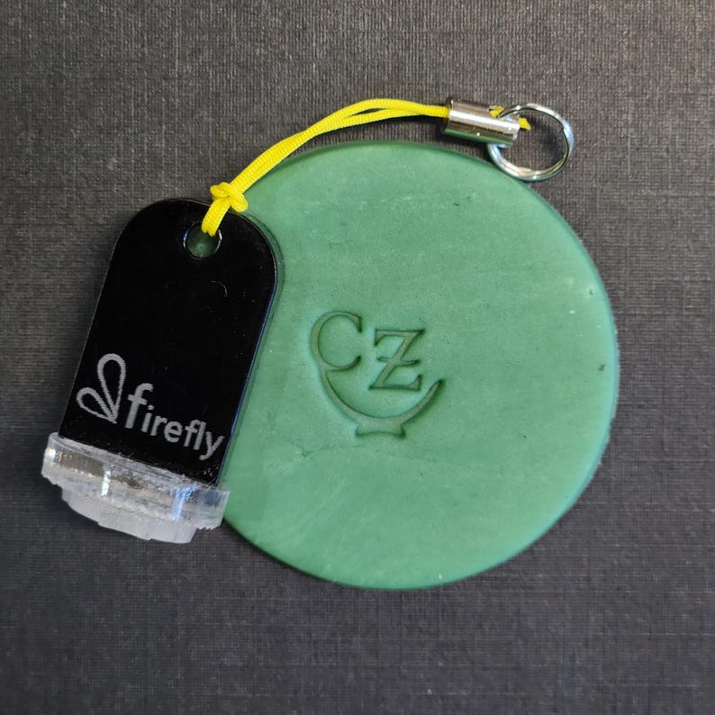 May include: A round, matte green disc featuring an embossed CZ logo, attached by a yellow cord to a black rectangular tag with the white text firefly and a stylized leaf icon, finished with a silver metal split ring for attachment.