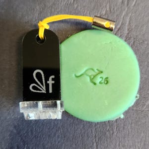 May include: A black tag with the letters "sf" in white, attached to a green circular object with a kangaroo graphic and the number "26". The tag has a clear plastic base and a yellow cord.