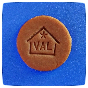 May include: A circular, light brown biscuit featuring the word "VAL" within a house design, with a star above. It rests on a square, textured blue surface. The biscuit's edges are slightly irregular.