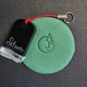 May include: A round, sage green clay disc featuring an embossed monogram of the letters A and B inside a circle. Attached to the disc is a black, rectangular Firefly brand stamp with a silver metal base and a red cord with a silver keyring.