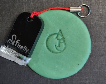 May include: A round, sage green clay disc featuring an embossed monogram of the letters A and B inside a circle. Attached to the disc is a black, rectangular Firefly brand stamp with a silver metal base and a red cord with a silver keyring.