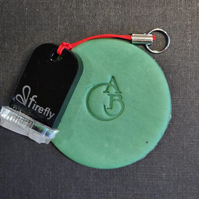 May include: A round, sage green clay disc featuring an embossed monogram of the letters A and B inside a circle. Attached to the disc is a black, rectangular Firefly brand stamp with a silver metal base and a red cord with a silver keyring.