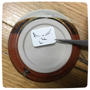 May include: A close-up of a ceramic plate with a brown and white glaze. A small white sticker with a black bird design is attached to the plate.