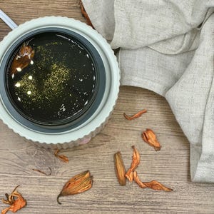 May include: A white and gray wax warmer with a dark liquid and gold flecks. Dried orange flower petals are scattered around the warmer on a wooden surface. A beige linen cloth is draped in the background.