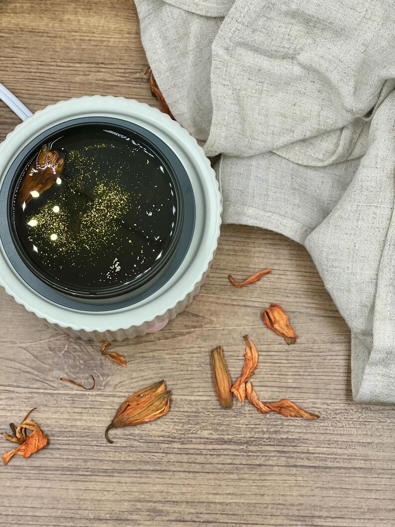 May include: A white and gray wax warmer with a dark liquid and gold flecks. Dried orange flower petals are scattered around the warmer on a wooden surface. A beige linen cloth is draped in the background.