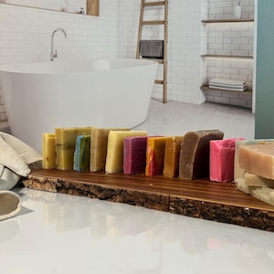 May include: A wooden board with a variety of colorful handmade soap bars. The soap bars are arranged in a row on the board, with the colors ranging from yellow to pink to purple. The board is sitting on a white surface.