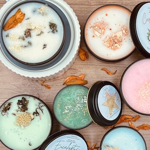 May include: A collection of scented soy wax candles in black tins, with various colors and decorative elements. Some candles have gold flakes, dried flowers, and botanical accents. The labels read "Eucalyptus Grove" and "Star Shine".