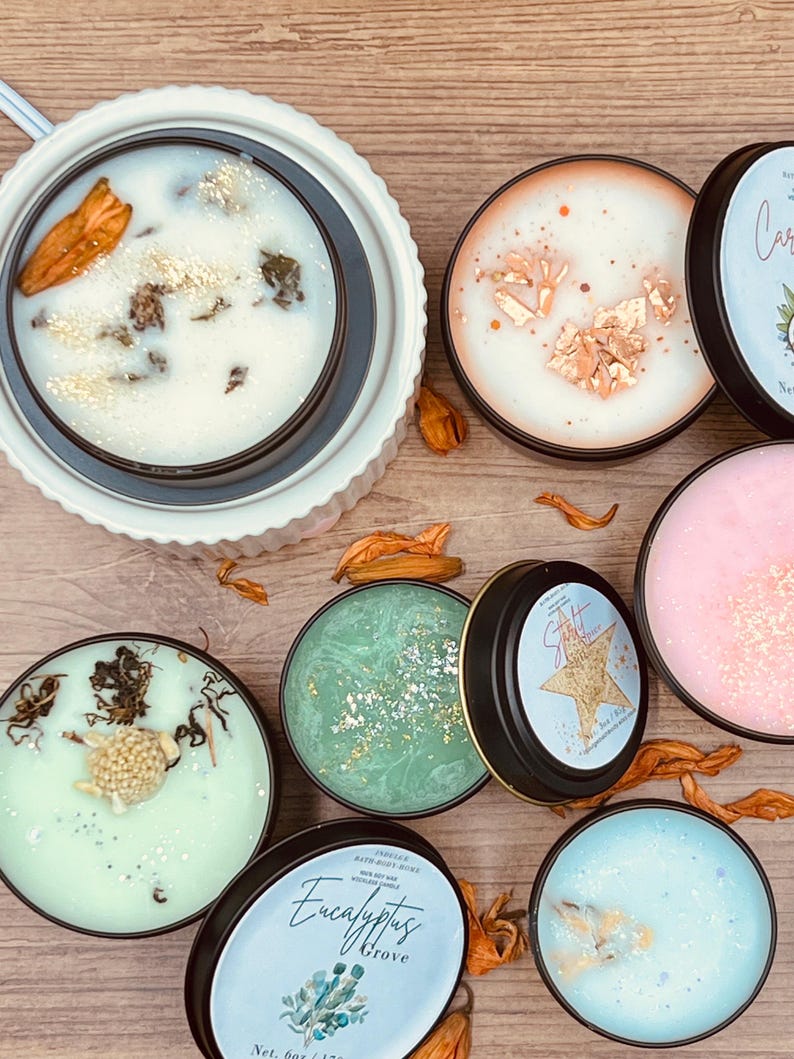 May include: A collection of scented soy wax candles in black tins, with various colors and decorative elements. Some candles have gold flakes, dried flowers, and botanical accents. The labels read "Eucalyptus Grove" and "Star Shine".