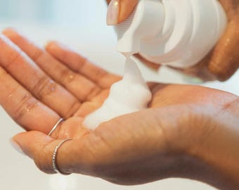 May include: A white pump bottle dispensing white foam into an open hand. The hand is adorned with a silver ring. The background is a blurred white surface. The foam appears to be a skincare product.