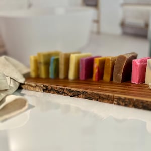 May include: A wooden tray with a natural bark edge holds a row of colorful handmade soap bars. The soaps are arranged in a rainbow pattern, with shades of green, blue, yellow, orange, red, and pink.