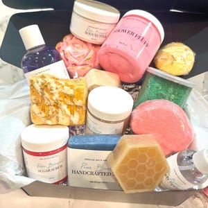 May include: A gift basket filled with bath and body products. The basket includes a pink shower fluff, a white body butter, a purple body wash, a pink sugar scrub, a honey-colored soap, a blue soap bar, a green bath bomb, a white bath bomb, a yellow bath bomb, and a clear body spray. The basket is lined with white tissue paper.