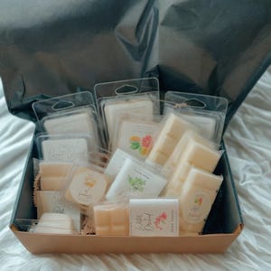 May include: A gift box filled with various wax melts in clear plastic packaging. The wax melts are in different shapes and sizes, with some labeled "Water Orchid" and "Wax Melts". The box is lined with black tissue paper.