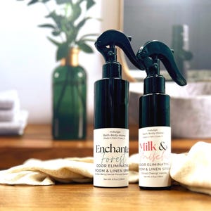 May include: Two dark green spray bottles labeled "Enchanted Forest" and "Milk & Mischief" for room and linen. The bottles are on a wooden surface with a cream-colored cloth. A green glass bottle and white towels are in the background.