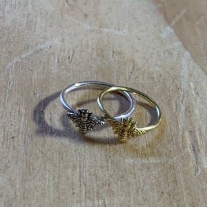 K&ouml;nnte beinhalten: Zwei Ringe mit Bienenmotiven. Ein Ring ist silberfarben, der andere goldfarben. Die Details der Bienen sind deutlich zu erkennen. Die Ringe liegen auf einer Holzoberfl&auml;che.