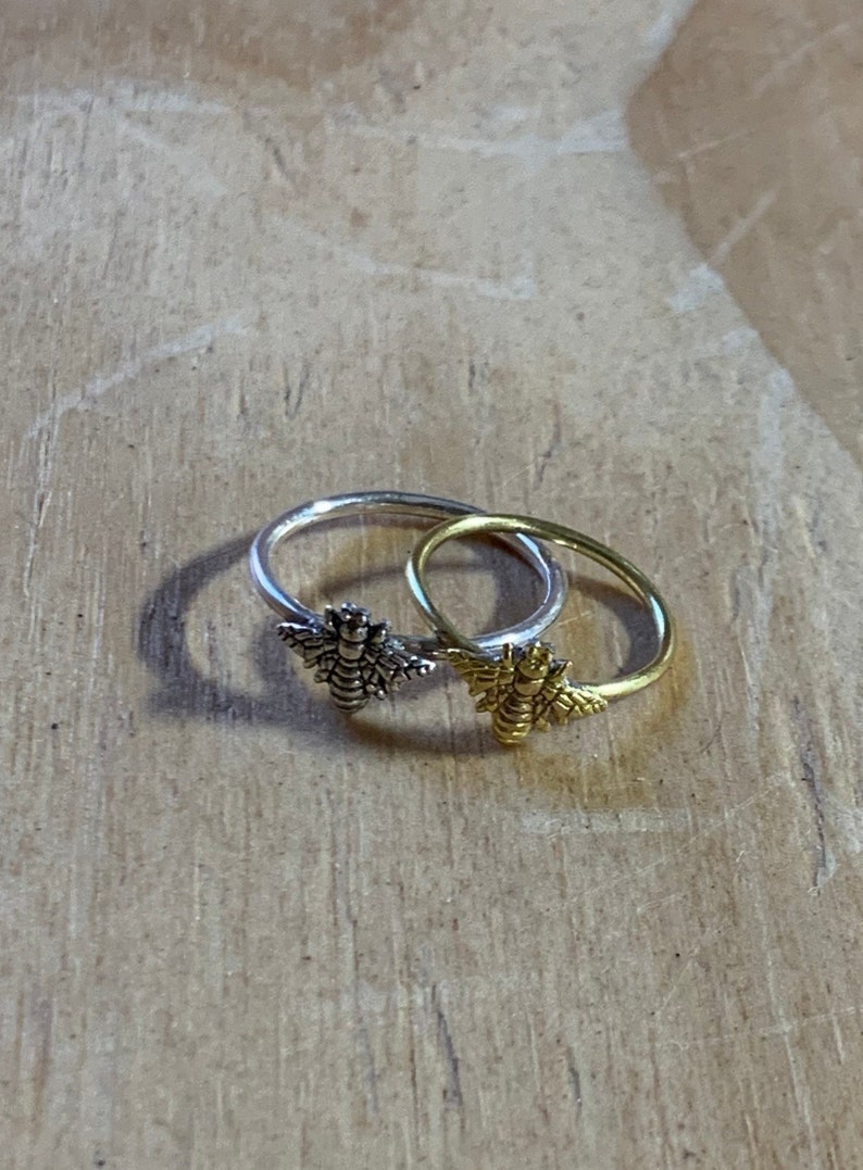 K&ouml;nnte beinhalten: Zwei Ringe mit Bienenmotiven. Ein Ring ist silberfarben, der andere goldfarben. Die Details der Bienen sind deutlich zu erkennen. Die Ringe liegen auf einer Holzoberfl&auml;che.