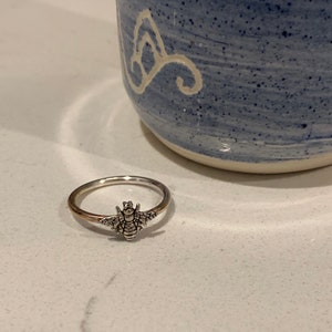 K&ouml;nnte beinhalten: Ein silberner Ring mit einem detaillierten Bienen-Design. Der Ring hat ein goldenes Band und einen silbernen Bienen-Anh&auml;nger mit ausgebreiteten Fl&uuml;geln. Er liegt auf einer wei&szlig;en Oberfl&auml;che, mit einer blau-wei&szlig;en Keramiktasse im Hintergrund.