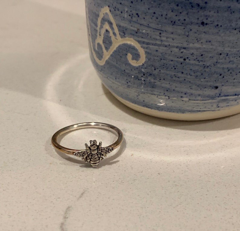 K&ouml;nnte beinhalten: Ein silberner Ring mit einem detaillierten Bienen-Design. Der Ring hat ein goldenes Band und einen silbernen Bienen-Anh&auml;nger mit ausgebreiteten Fl&uuml;geln. Er liegt auf einer wei&szlig;en Oberfl&auml;che, mit einer blau-wei&szlig;en Keramiktasse im Hintergrund.