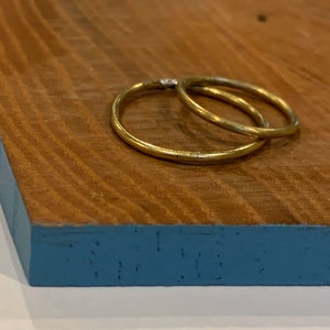 Puede incluir: Dos anillos dorados, uno ligeramente m&aacute;s grande que el otro, entrelazados. Los anillos descansan sobre una superficie de madera con un borde pintado de azul. La veta de la madera es visible, agregando textura a la imagen.