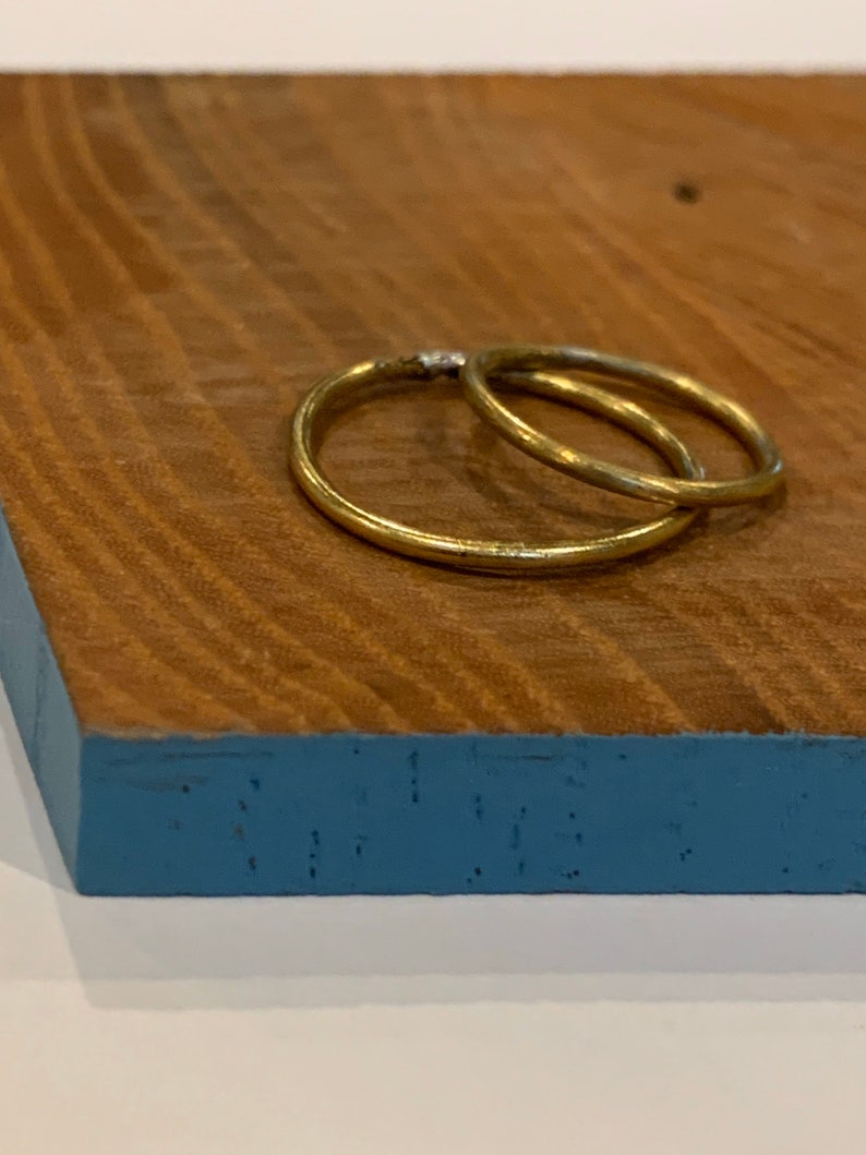 Puede incluir: Dos anillos dorados, uno ligeramente m&aacute;s grande que el otro, entrelazados. Los anillos descansan sobre una superficie de madera con un borde pintado de azul. La veta de la madera es visible, agregando textura a la imagen.