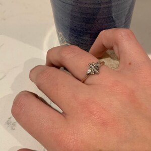 K&ouml;nnte beinhalten: Silberring mit detailliertem floralem Design. Der Ring befindet sich an einem Finger, mit einer blauen Keramiktasse im Hintergrund. Die filigranen Details des Rings sind sichtbar und zeigen eine zarte, von der Natur inspirierte &Auml;sthetik.