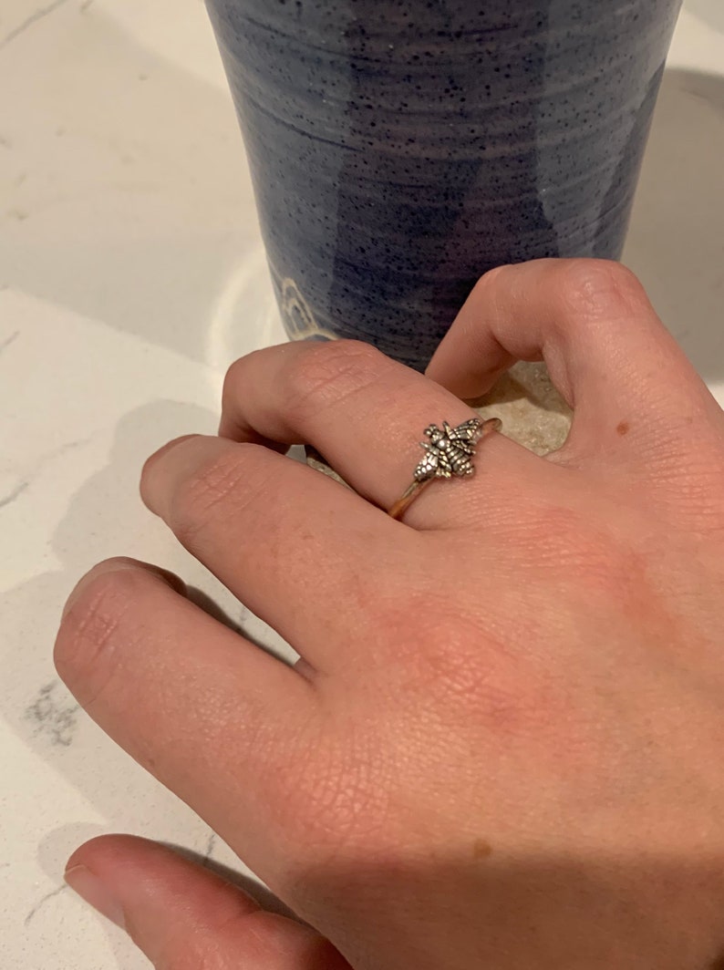 K&ouml;nnte beinhalten: Silberring mit detailliertem floralem Design. Der Ring befindet sich an einem Finger, mit einer blauen Keramiktasse im Hintergrund. Die filigranen Details des Rings sind sichtbar und zeigen eine zarte, von der Natur inspirierte &Auml;sthetik.