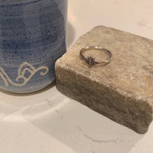 K&ouml;nnte beinhalten: Ein silberner Ring mit einem Bienenmotiv, pr&auml;sentiert auf einem hellen Stein. Im Hintergrund ist ein blauer Keramikbecher zu sehen. Der Ring zeigt eine detaillierte Biene. Ein filigranes Schmuckst&uuml;ck.
