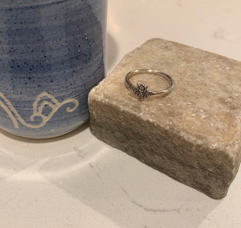 K&ouml;nnte beinhalten: Ein silberner Ring mit einem Bienenmotiv, pr&auml;sentiert auf einem hellen Stein. Im Hintergrund ist ein blauer Keramikbecher zu sehen. Der Ring zeigt eine detaillierte Biene. Ein filigranes Schmuckst&uuml;ck.