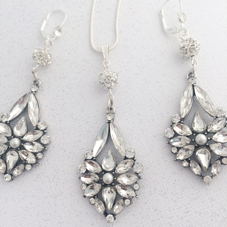 May include: A silver necklace and earrings set with crystal teardrop pendants. The pendants are adorned with clear rhinestones and a single pearl in the center.
