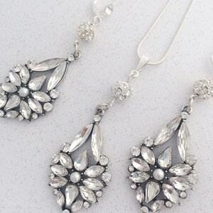 May include: A silver necklace and earrings set with crystal teardrop pendants. The necklace has a delicate chain and a small crystal ball charm. The earrings have a clip-on closure.