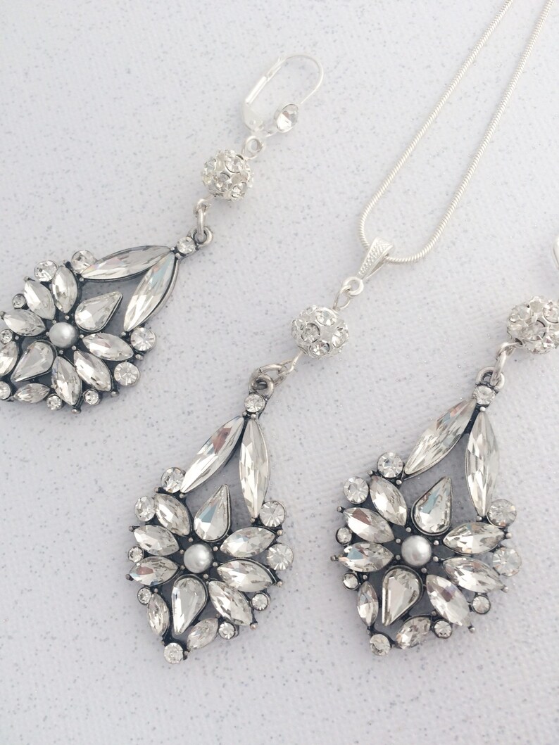 May include: A silver necklace and earrings set with crystal teardrop pendants. The necklace has a delicate chain and a small crystal ball charm. The earrings have a clip-on closure.